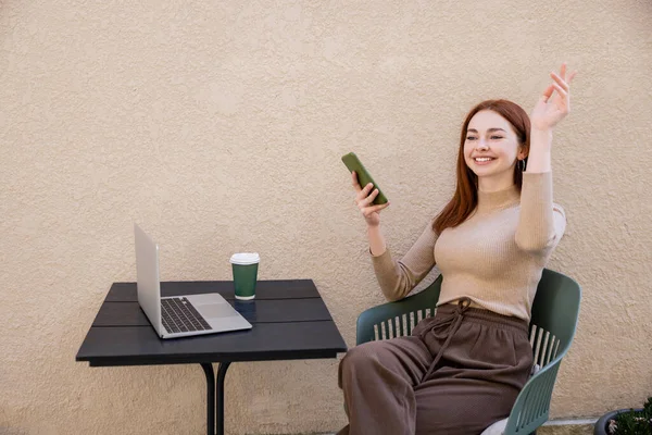 Redhead freelancer in turtleneck holding smartphone while calling waiter on summer terrace — Stock Photo