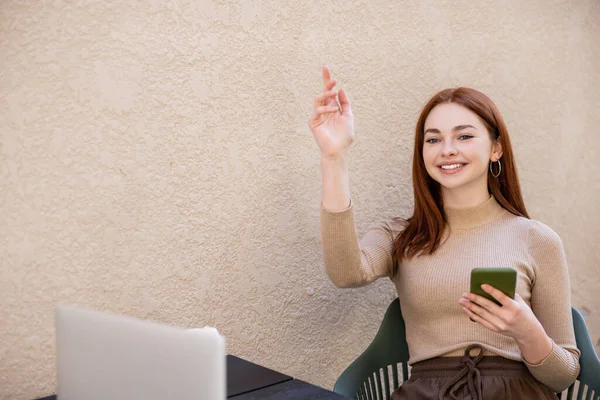 Glückliche Freiberuflerin im Rollkragen hält Smartphone in der Hand, während sie Kellner auf Sommerterrasse anruft — Stockfoto