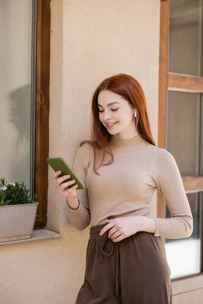 Happy redhead woman in wireless earphone holding smartphone and listening music - foto de stock
