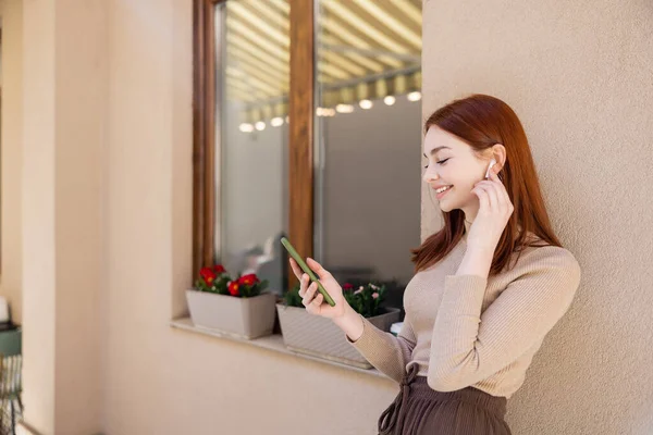 Mulher ruiva satisfeito ajustando fone de ouvido sem fio e segurando smartphone enquanto escuta música — Fotografia de Stock