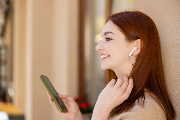 Visão lateral da mulher positiva no fone de ouvido sem fio segurando smartphone e ouvindo música — Fotografia de Stock