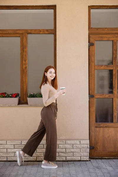 Full length of happy young woman in wireless earphone holding paper cup and walking on street — стоковое фото