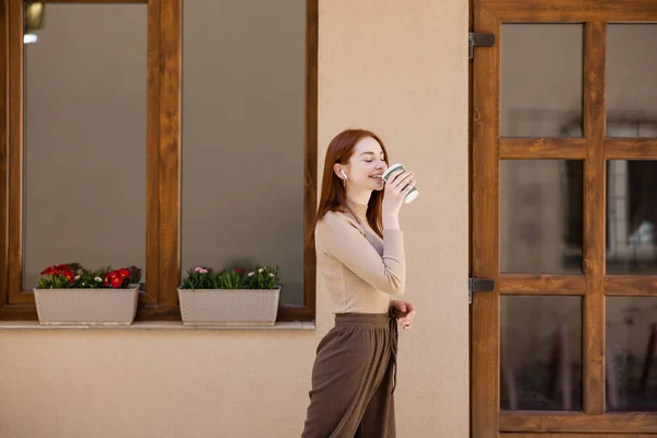 Heureuse jeune femme dans écouteur sans fil boire du café pour aller — Photo de stock