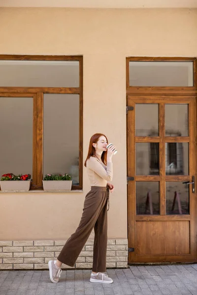 Full length of stylish redhead woman in earphone drinking coffee to go — стоковое фото