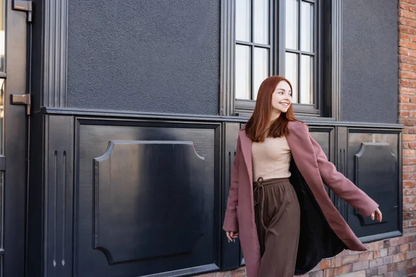 Glückliche rothaarige Frau in stylischem Mantel, die in der Nähe moderner Gebäude spaziert — Stockfoto