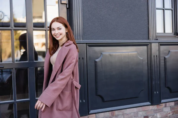 Glückliche rothaarige Frau in stylischem Mantel lächelt in der Nähe moderner Gebäude — Stockfoto