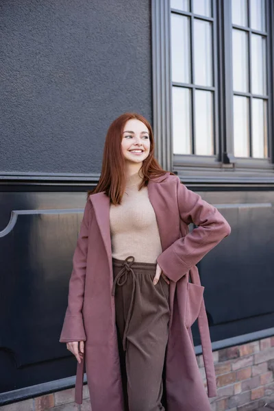 Heureuse rousse femme en manteau élégant debout avec la main sur la hanche près du bâtiment moderne — Photo de stock