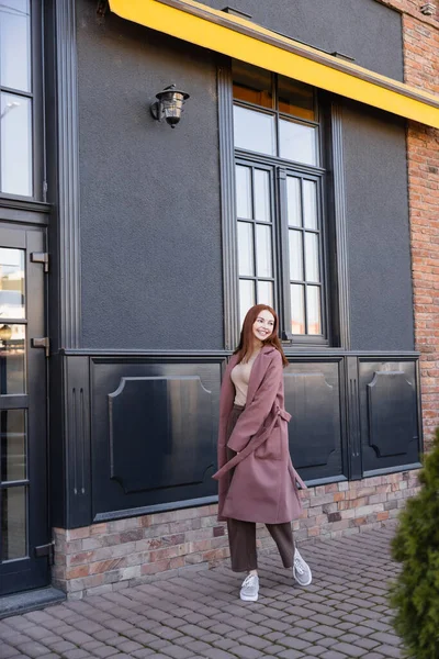 Pleine longueur de femme heureuse en manteau élégant marchant près du bâtiment moderne — Photo de stock