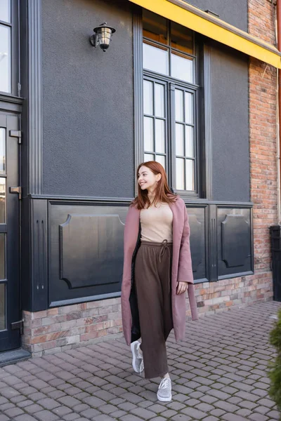 Longitud completa de mujer pelirroja sonriente en abrigo elegante caminando cerca de edificio moderno - foto de stock