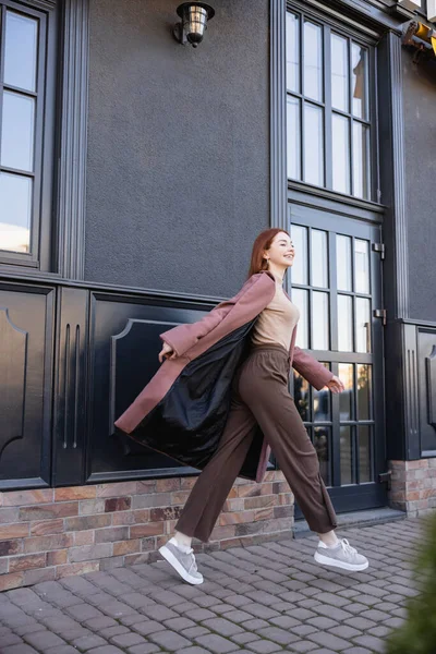 Повна довжина радісної рудої жінки в стильному пальто, що йде біля сучасної будівлі — стокове фото