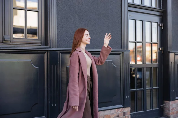 Happy redhead woman in stylish coat waving hand near modern building — Stock Photo