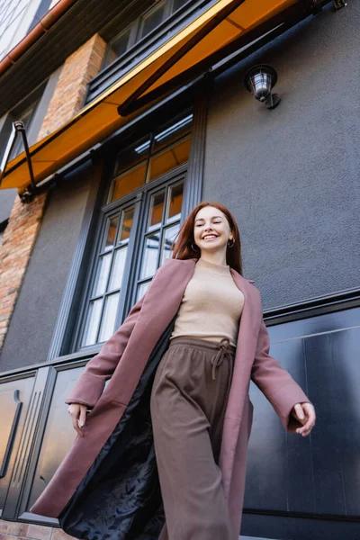 Vue à angle bas de femme rousse heureuse en manteau élégant marchant près du bâtiment moderne — Photo de stock