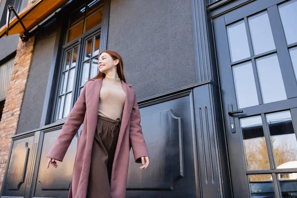 Vista de ángulo bajo de la mujer pelirroja complacida en abrigo elegante caminando cerca de edificio moderno - foto de stock