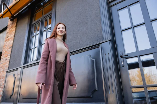 Blick auf eine junge rothaarige Frau im eleganten Mantel, die lächelt und in der Nähe moderner Gebäude spaziert — Stockfoto