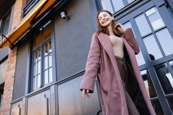 Low angle view of positive and redhead woman in stylish coat walking near modern building — стоковое фото