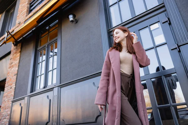 Vue à angle bas de la femme heureuse dans un manteau élégant ajustant les cheveux rouges près du bâtiment moderne — Photo de stock