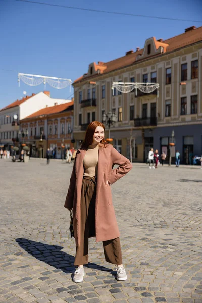 Full length of happy woman with red hair posing with hand on hip on european street — Photo de stock