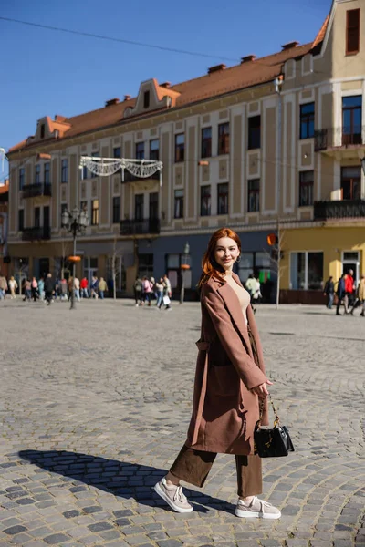 Full length of pleased woman with red hair holding handbag on european street — Photo de stock