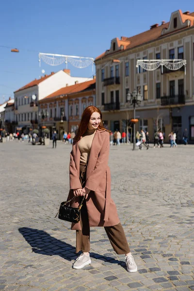 Full length of happy woman with red hair holding handbag on european street — Photo de stock