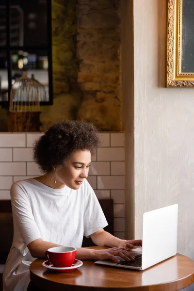 Young african american woman using laptop near cup of coffee in cafe — Stock Photo