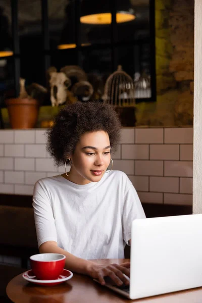 Young african american woman using laptop near cup of coffee on table — Stock Photo