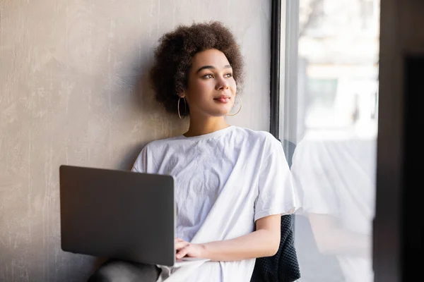 Young african american woman in white t-shirt holding laptop near window — Stock Photo