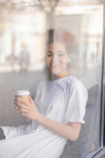 Happy african american woman in white t-shirt holding paper cup behind window — Stock Photo