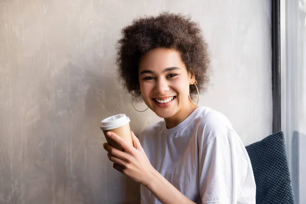 Joyful african american woman in white t-shirt holding paper cup — Stock Photo