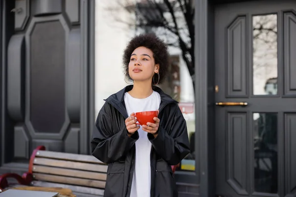Mujer afroamericana en chaqueta sosteniendo taza de café en la calle - foto de stock
