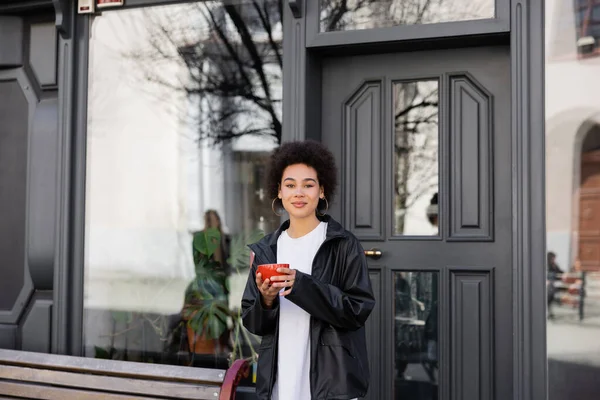 Pleased african american woman in jacket holding cup of coffee on street — Stock Photo