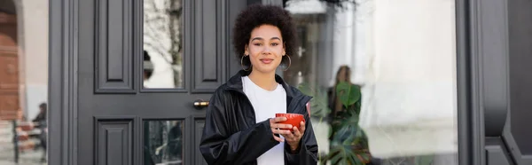 Pleased african american woman in jacket holding cup of coffee on street, banner — Stockfoto