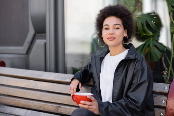 Jeune femme afro-américaine en veste tenant une tasse de café et assise sur un banc — Photo de stock