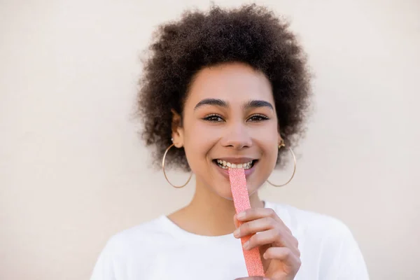 Joyful african american young woman eating sweet jelly strip on white - foto de stock