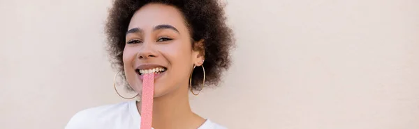 Happy african american young woman eating sweet jelly bubble tape on white, banner - foto de stock