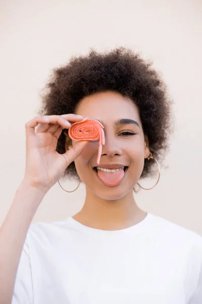 Feliz africano americano joven mujer cubriendo ojo con gelatina burbuja cinta y sobresaliendo lengua en blanco — Stock Photo