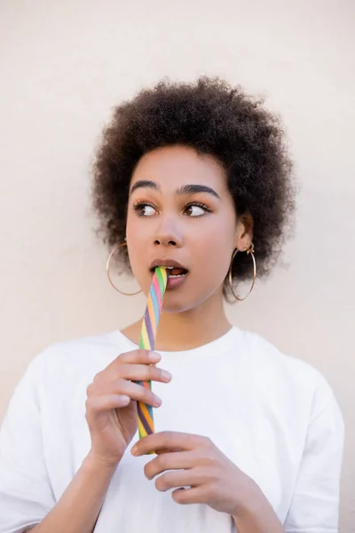 African american young woman in hoop earrings eating jelly strip on white — Stockfoto