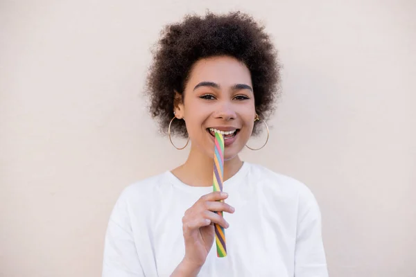Happy african american young woman in hoop earrings eating jelly strip on white — Fotografia de Stock