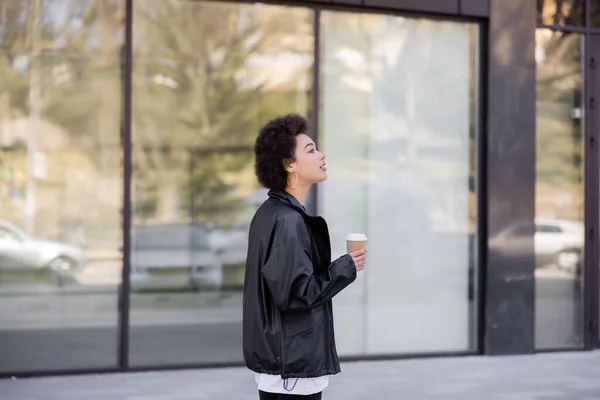 Frisé afro-américaine femme en veste tenant tasse en papier à l'extérieur — Photo de stock