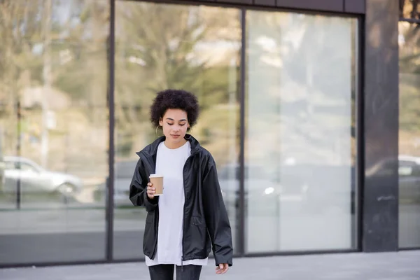 Curly african american woman in jacket holding paper cup on urban street — Fotografia de Stock