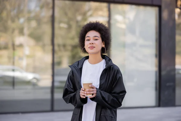 Young african american woman in jacket holding paper cup on urban street — Stock Photo