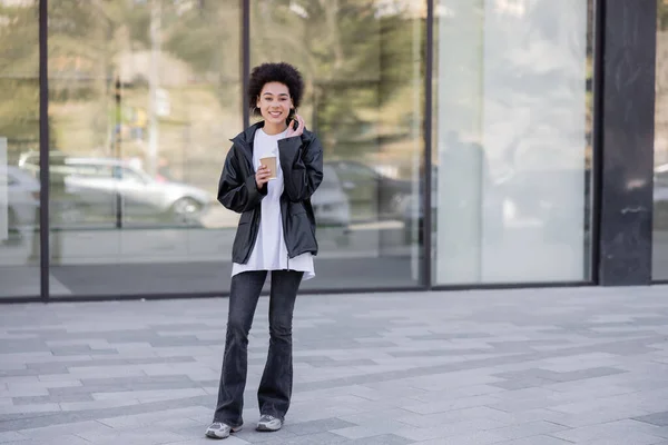 Comprimento total de mulher americana africana alegre na jaqueta segurando copo de papel na rua urbana — Fotografia de Stock