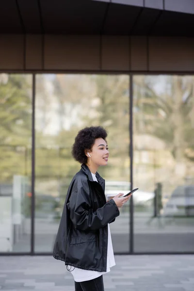 Cheerful african american woman in jacket and earphone holding smartphone outside — Foto stock