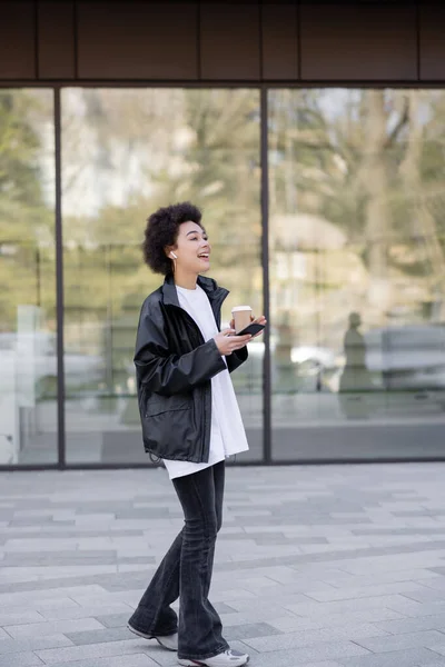 Full length of cheerful african american woman in earphone holding paper cup and smartphone outside — Stock Photo