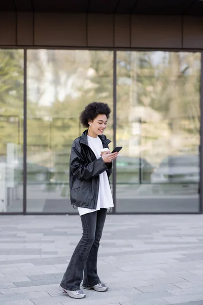 Pleine longueur de souriante femme afro-américaine dans un casque tenant tasse en papier et en utilisant un smartphone à l'extérieur — Photo de stock