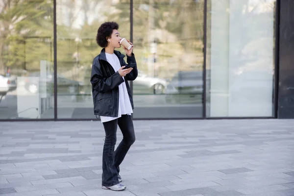 Full length of curly african american woman in earphone drinking coffee to go and holding smartphone outside — Stock Photo