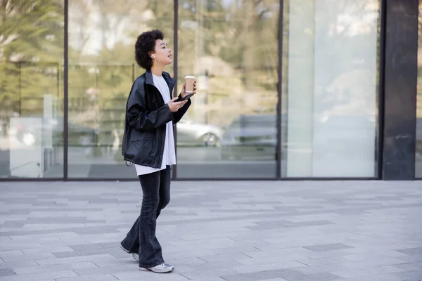 Full length of curly african american woman in earphone holding paper cup and smartphone outside — Stock Photo