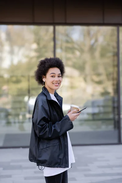 Smiling african american woman in earphone holding paper cup and smartphone outside — Photo de stock