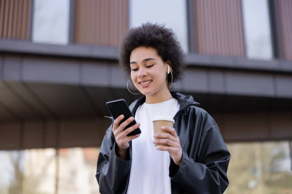 Vue à faible angle de souriante femme afro-américaine dans un écouteur tenant tasse en papier et smartphone à l'extérieur — Photo de stock