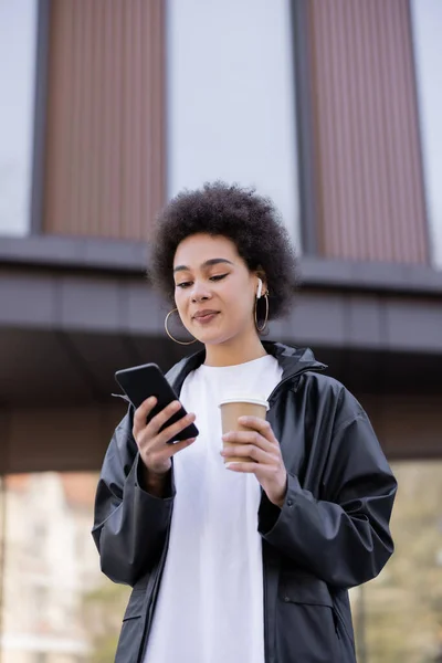 Low angle view of curly african american woman in earphone holding paper cup and smartphone outside — Stock Photo