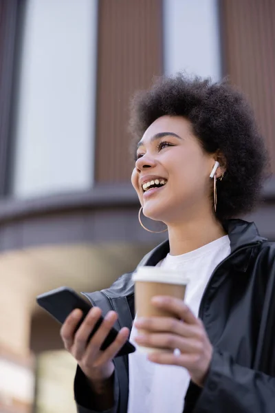 Vue à faible angle de la femme afro-américaine joyeuse dans un écouteur tenant tasse en papier et smartphone à l'extérieur — Photo de stock